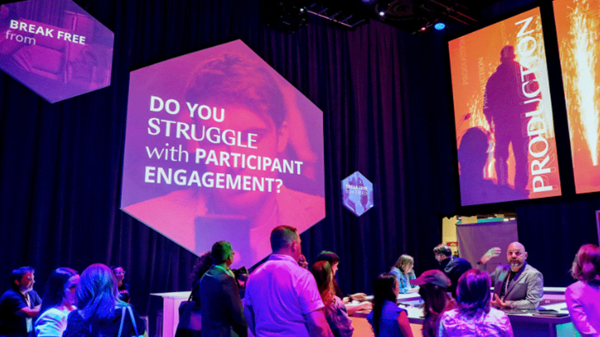 People gather at an event with large screens displaying messages, including “Do you struggle with participant engagement?” The environment is colorful with purple and blue lighting and geometric screen shapes.