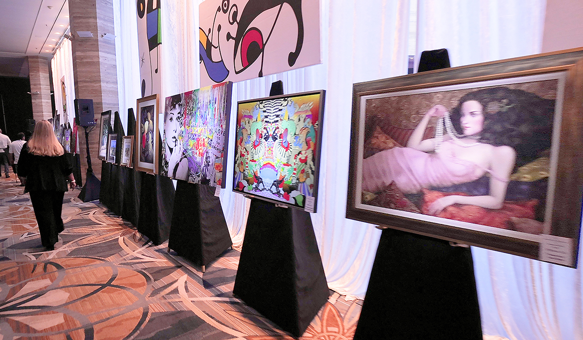 Several framed artworks are displayed on easels along a carpeted hallway at an indoor exhibition. A person with long hair walks past the art, and white drapes cover the windows in the background.
