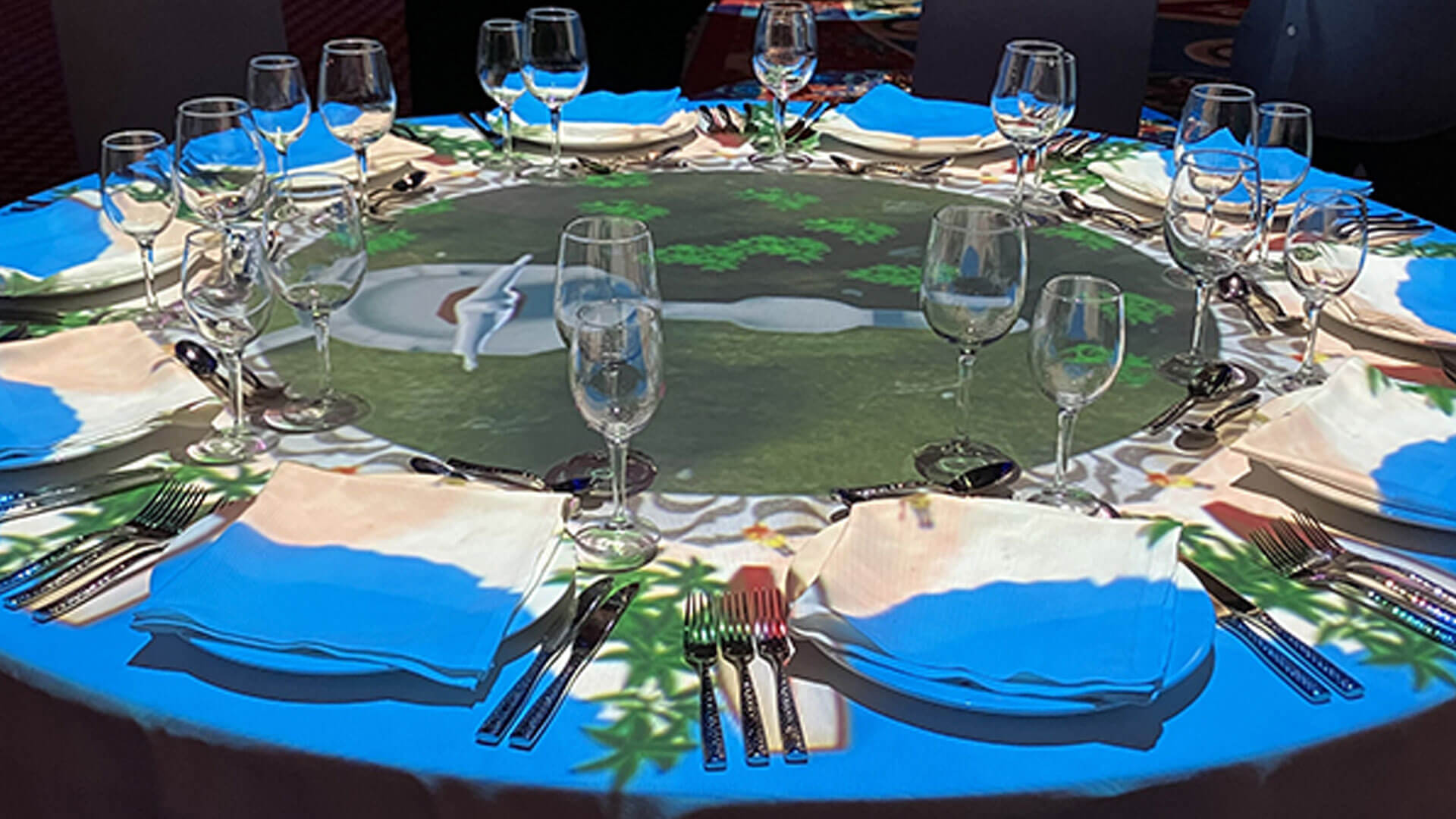 A round dining table set with glasses, plates, and cutlery, featuring a digital projection of a scenic landscape with blue skies, greenery, and a white path on the tablecloth.