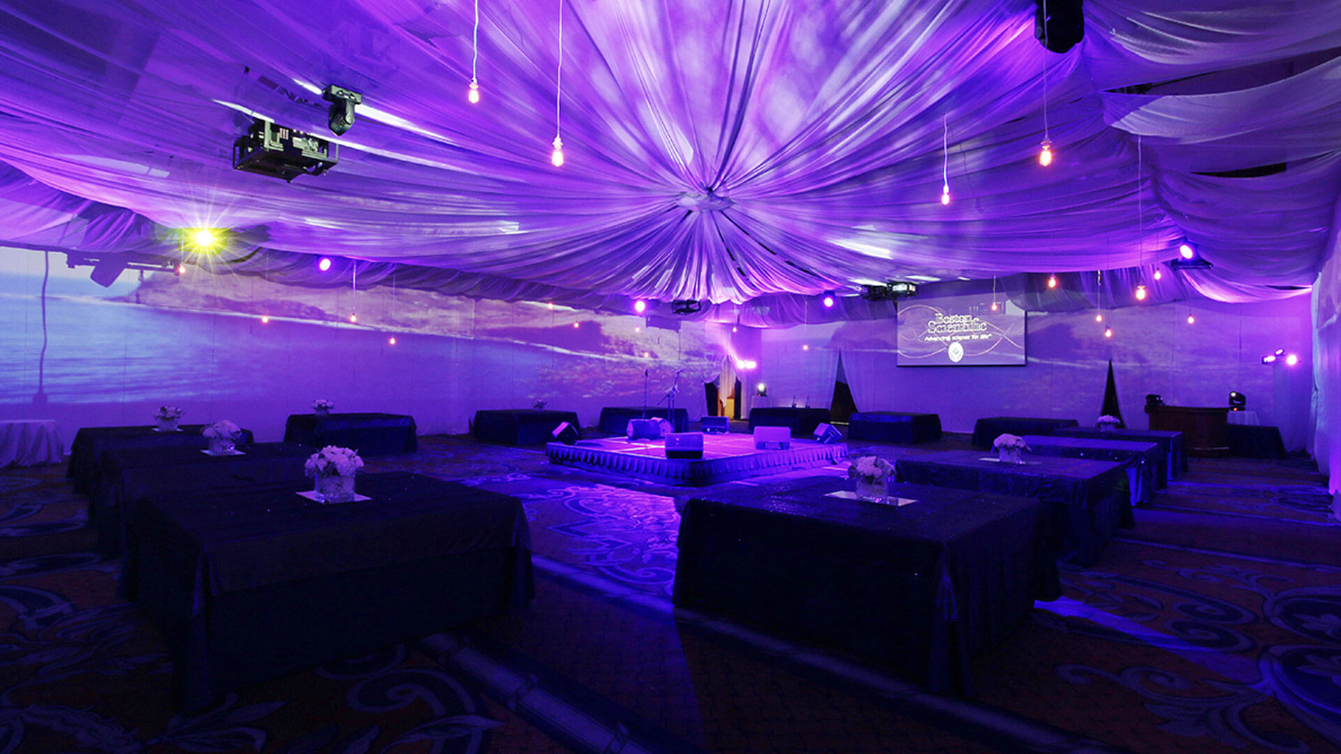 A large event hall decorated with draped white fabric and purple lighting, featuring low tables with floral centerpieces, a central stage, and projected images on the walls. The room is set up for a formal gathering.