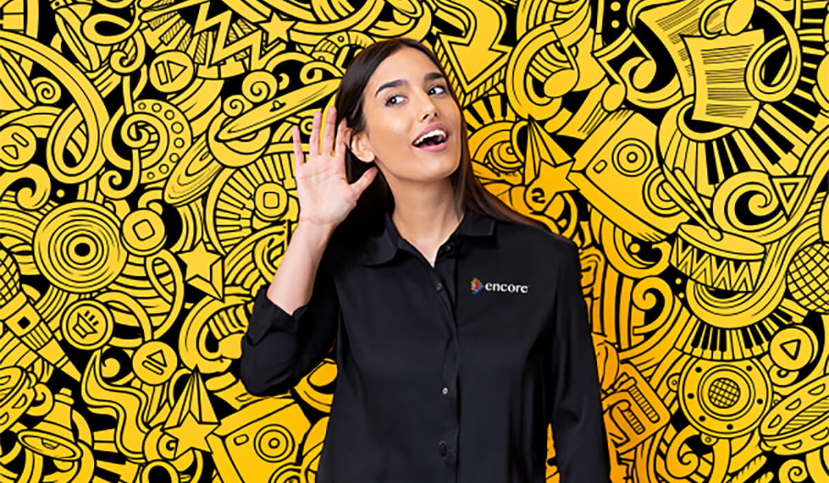 A woman wearing a black Encore shirt stands in front of a vibrant yellow and black abstract doodle mural, holding a hand to her ear as if trying to listen closely.