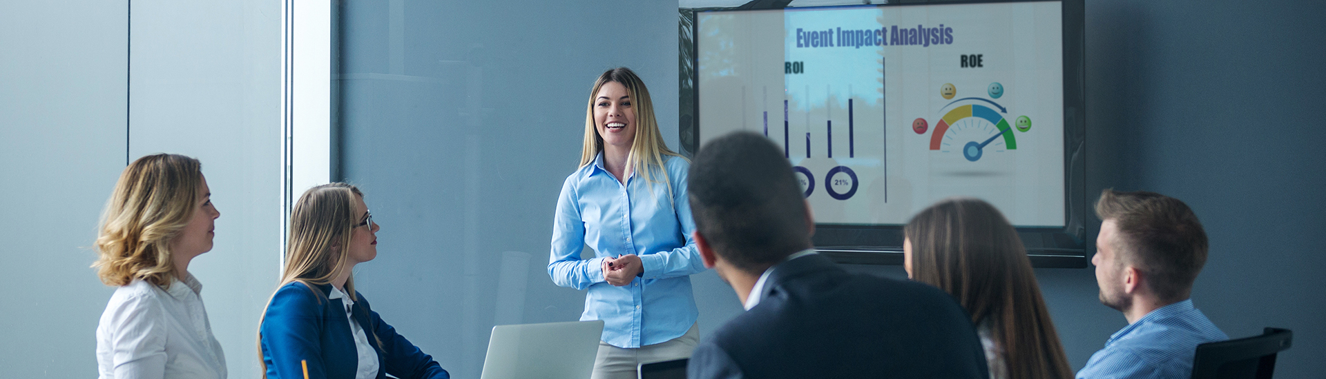 A woman stands and presents to five colleagues in a conference room, smiling. A large screen displays graphs and the text Event Impact Analysis with bar charts labeled ROI and ROE.