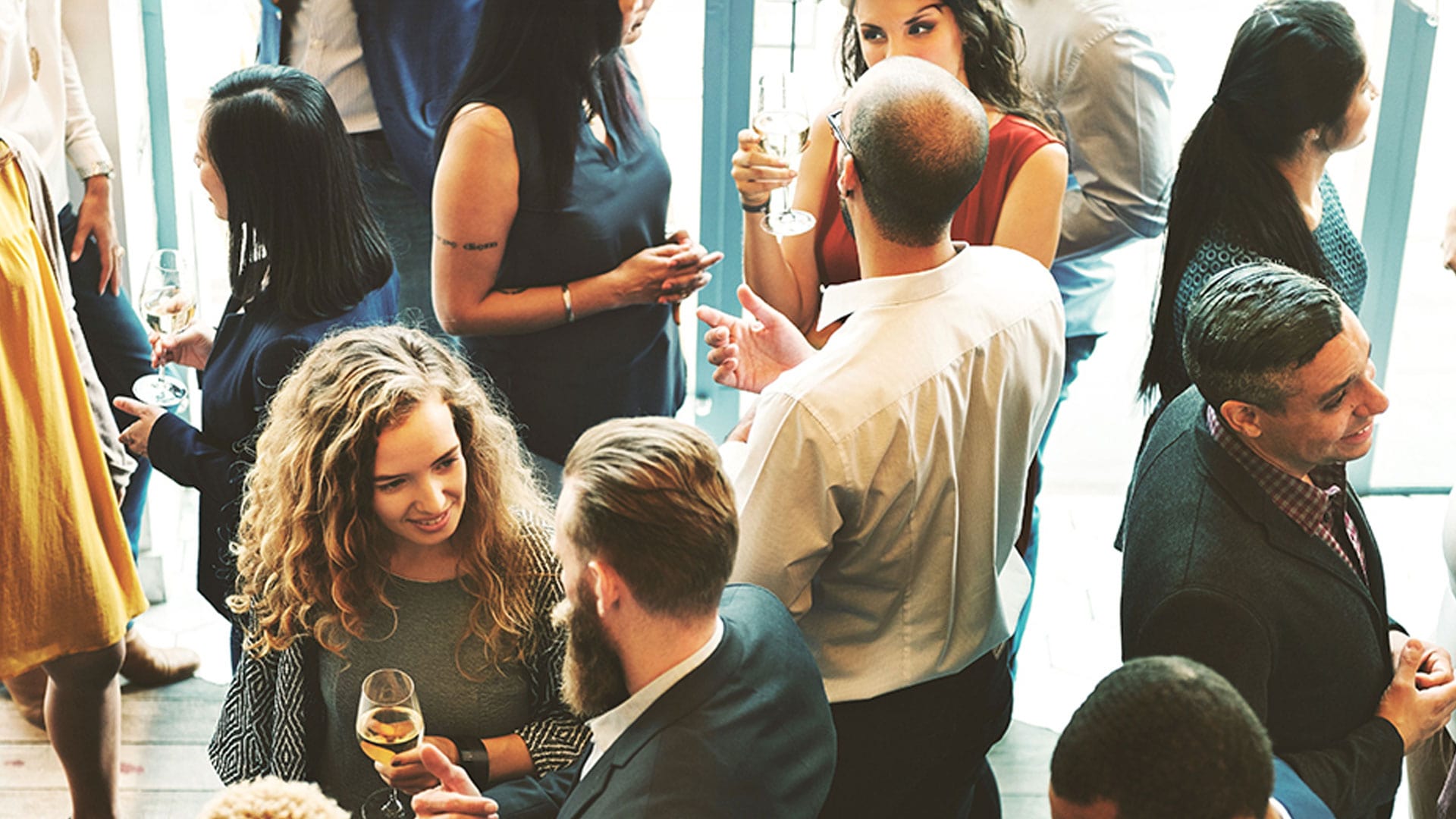 A group of people dressed in business attire stand close together at a social event, holding drinks, conversing, and smiling in a bright, modern indoor setting.