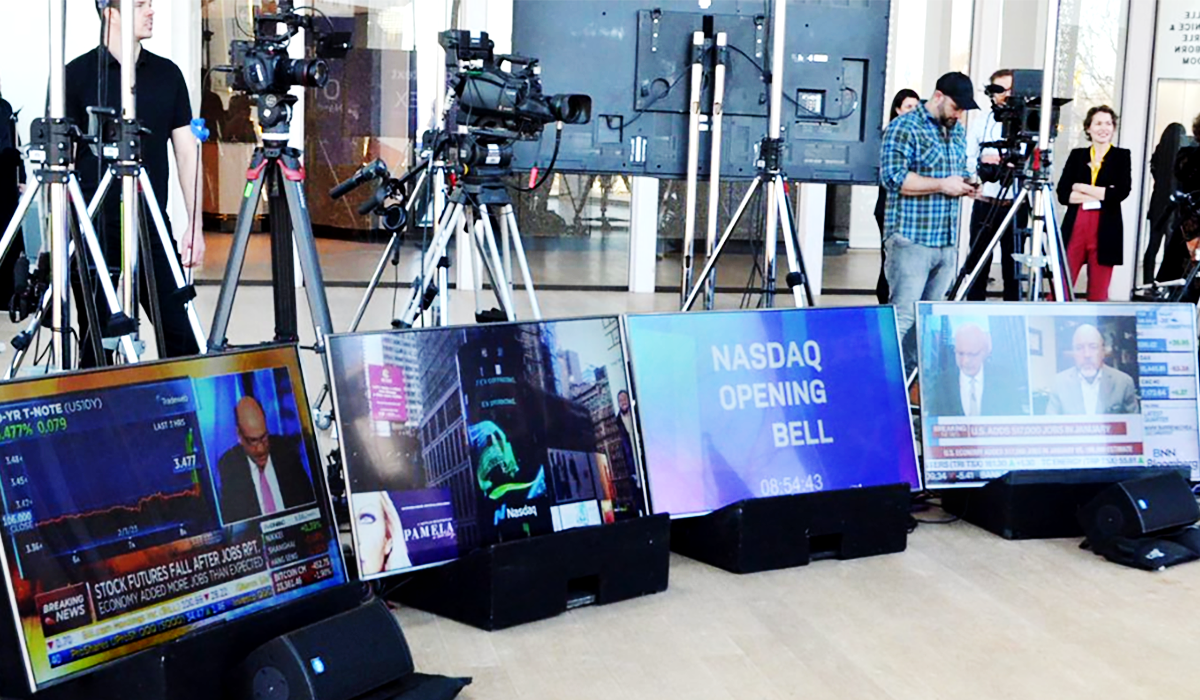 Several television cameras and crew film in a studio with large screens displaying live financial news, stock charts, and the text “NASDAQ OPENING BELL.” Reporters and staff stand in the background.
