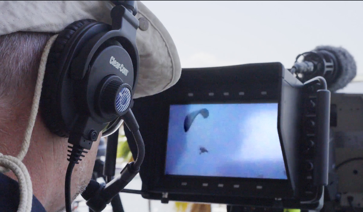 A person wearing headphones and a hat closely observes a monitor displaying footage of an unidentified flying object in the sky, with professional camera equipment visible.