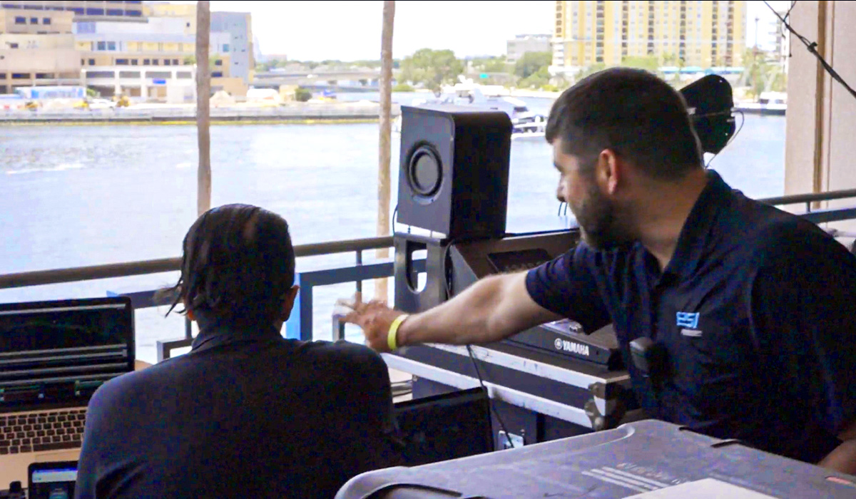 Two people working at an outdoor audio-visual setup; one points toward a laptop while the other looks on. Large speakers and equipment surround them, with a waterfront view in the background.