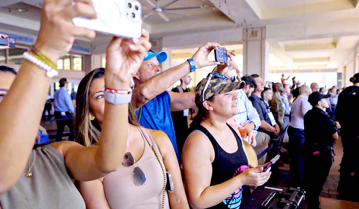 A crowd of people stands indoors, many holding up smartphones and cameras to take photos or videos. Some appear focused on the event, while others interact with their devices. Ceiling fans and natural light are visible in the background.