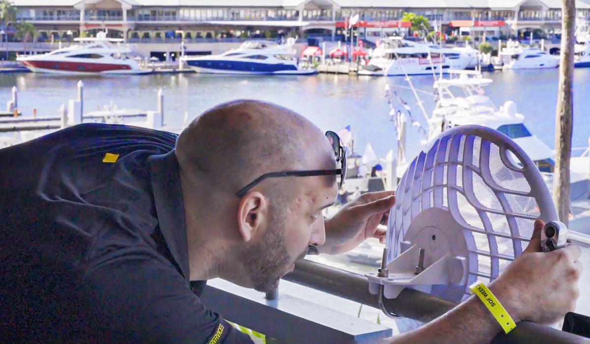 A man in a black shirt installs or adjusts a satellite dish on a balcony overlooking a marina filled with yachts and boats on a sunny day.