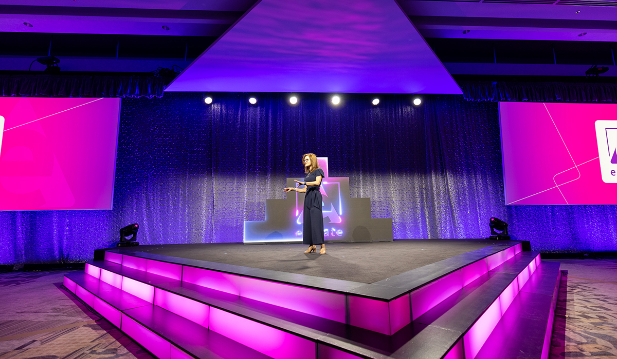 A woman in a navy dress stands alone on a brightly lit, triangular stage with pink and purple lighting, presenting at an event. Large display screens with geometric designs frame the background.