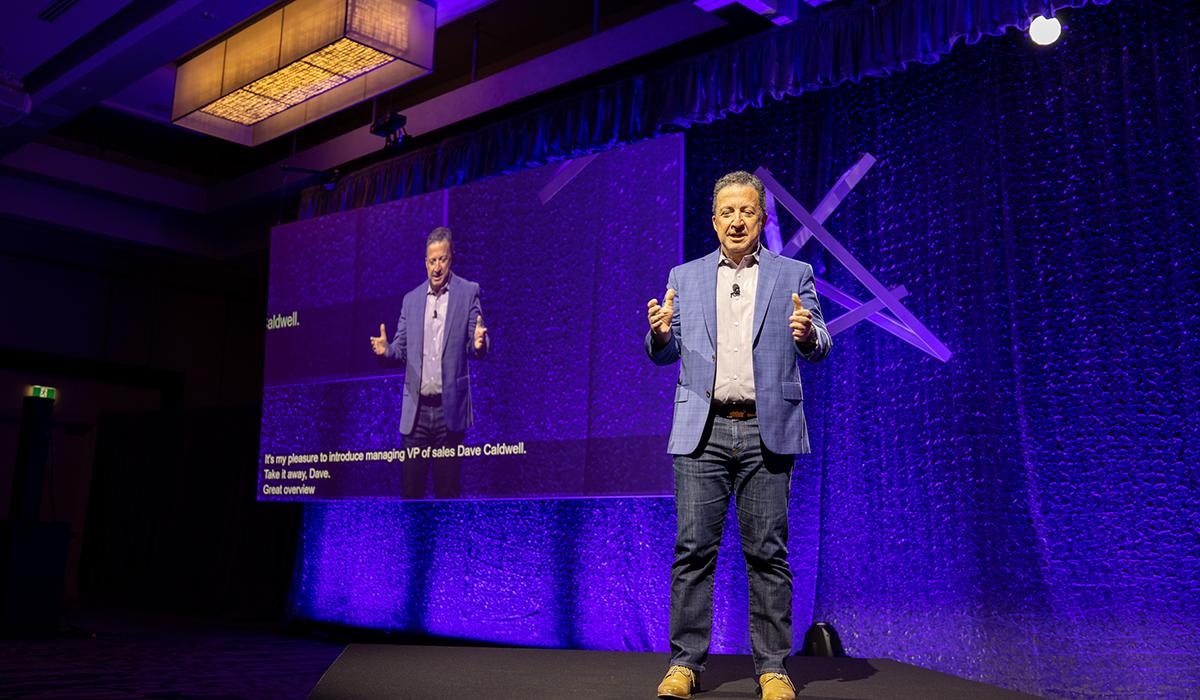 A man in a blue suit jacket and jeans stands on a stage, speaking to an audience. A large screen behind him displays his image and text. The background is lit with purple lighting.