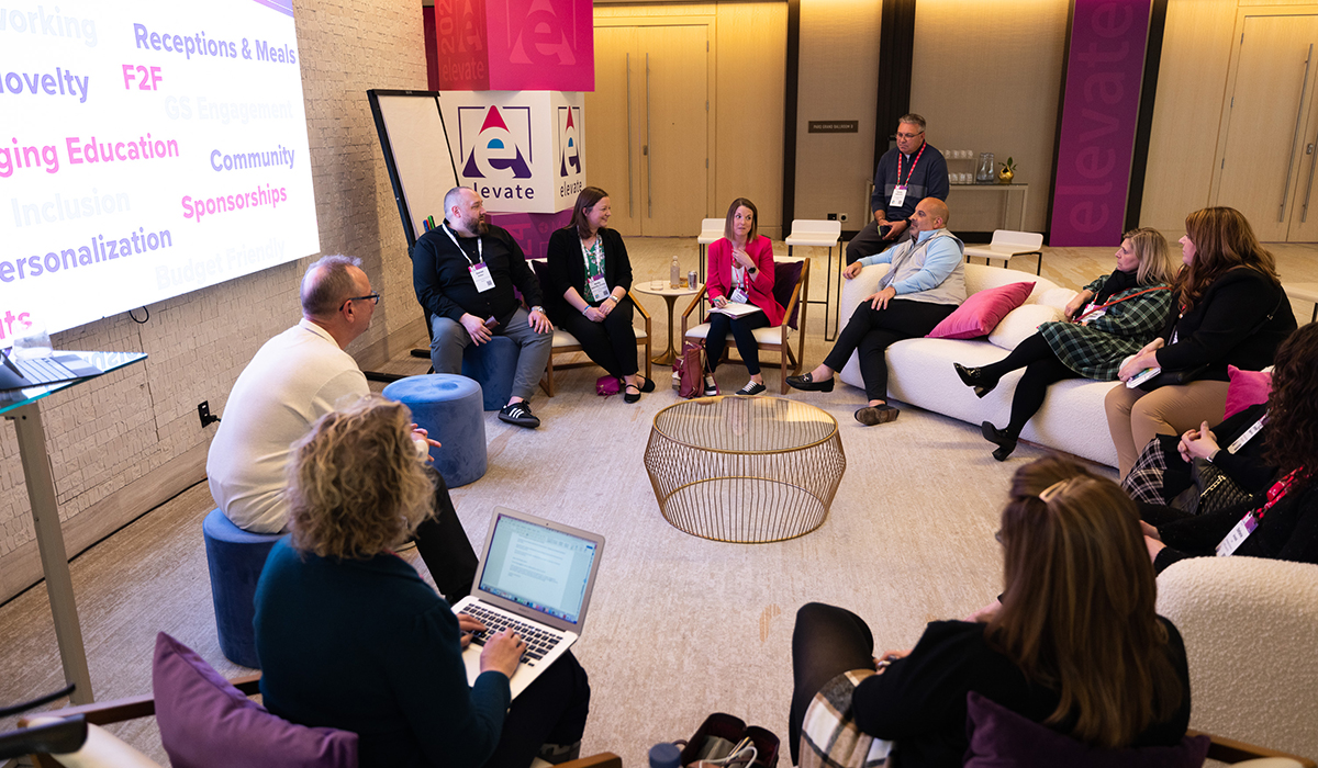 A group of people sits in a circle in a modern lounge, engaged in discussion. A presentation board and large screen with colorful text are visible in the background. One person is taking notes on a laptop in the foreground.