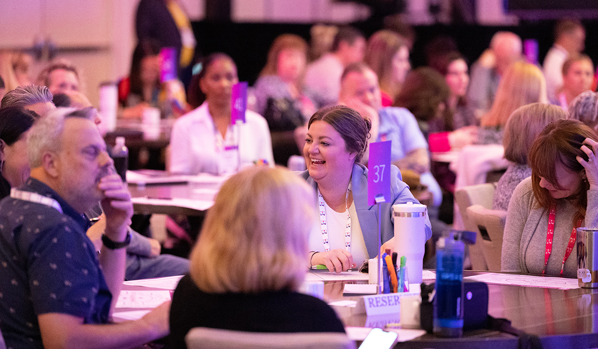 A group of people sit at round tables in a busy conference room, talking and smiling. One woman in the center is laughing, and there are papers, lanyards, and table number signs visible.