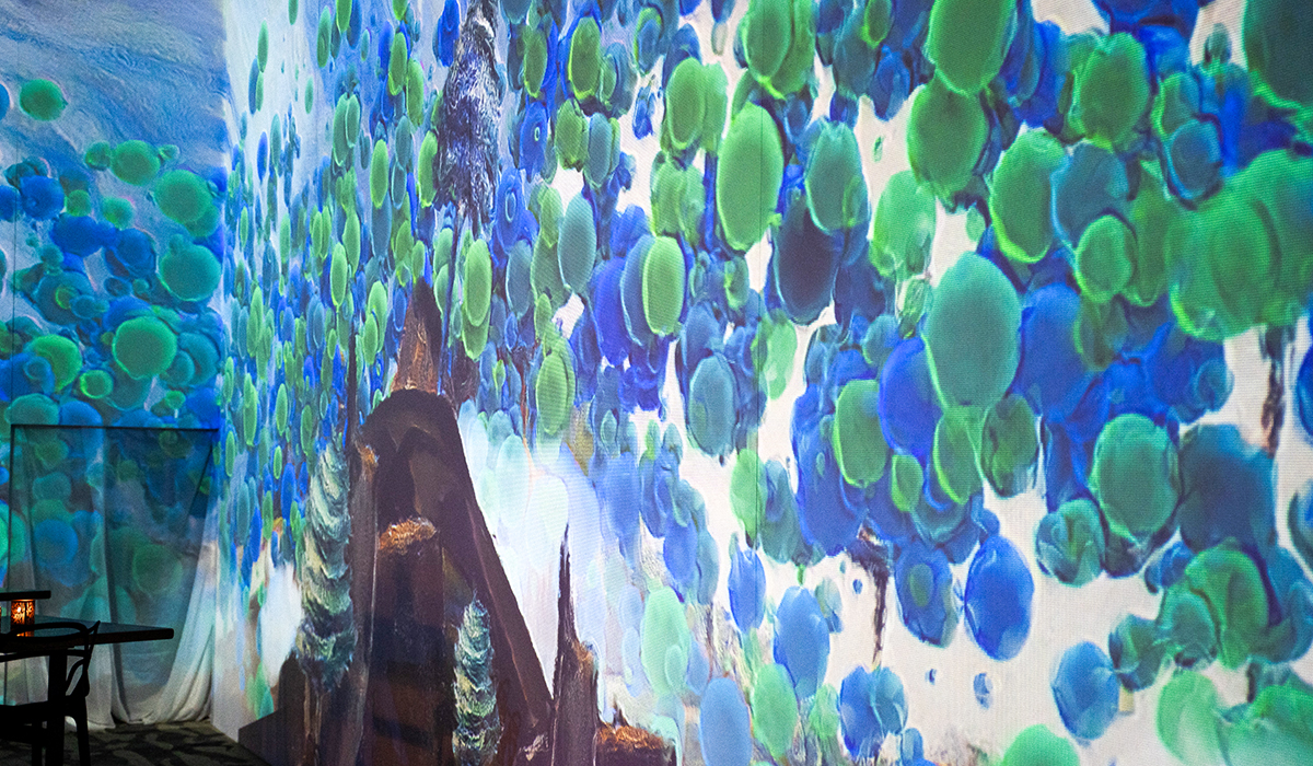 A colorful digital art projection covers a wall, featuring clusters of blue and green circular shapes with abstract scenery, creating an immersive visual experience. A small table with a lamp is visible in the corner.