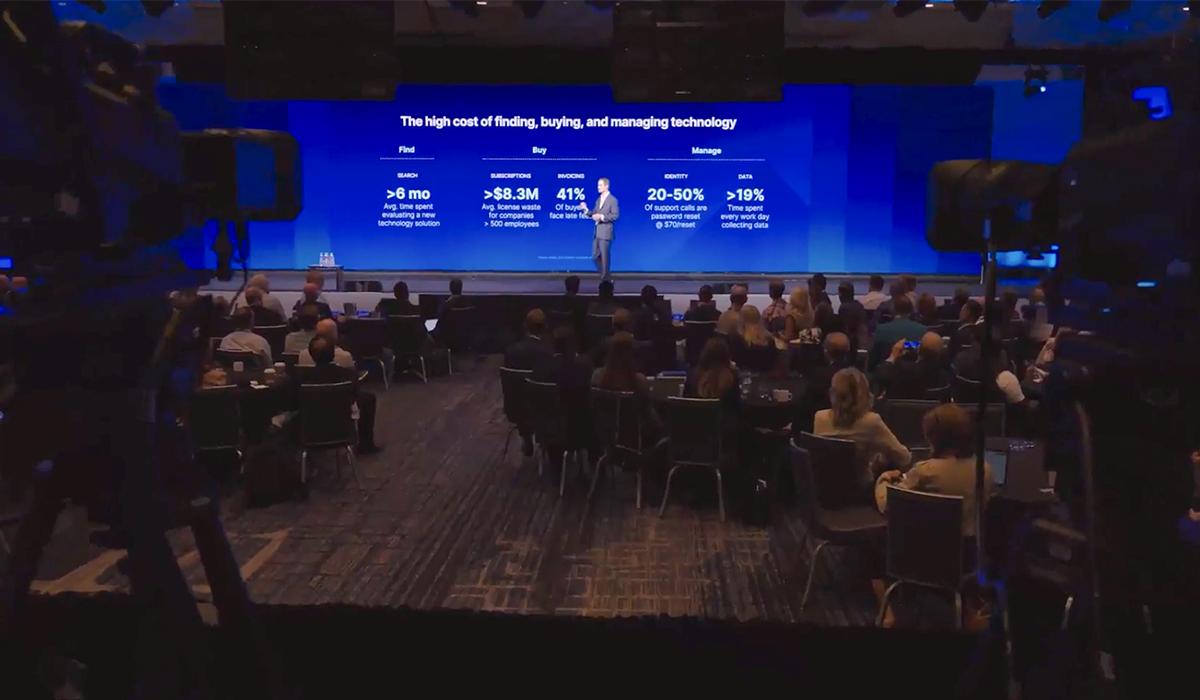 A speaker stands on stage presenting to an audience in a conference room. A large blue screen behind shows statistics and text about technology costs, with seated attendees facing the stage.