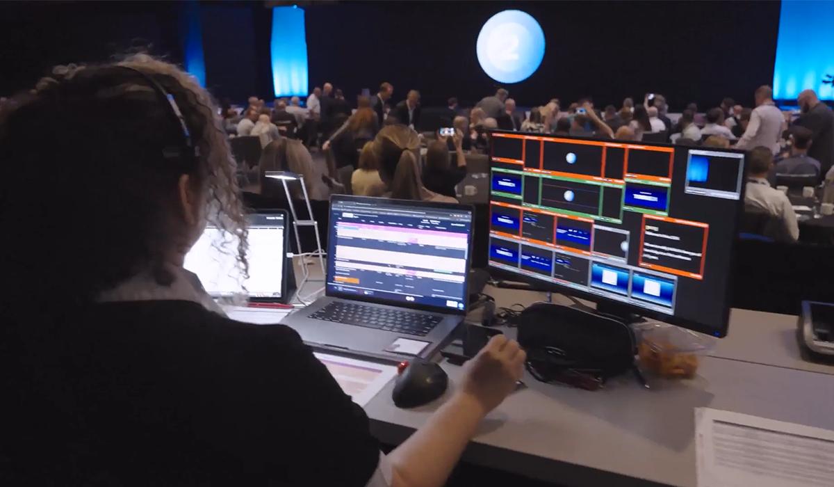 A person with a headset operates laptops and monitors displaying video feeds and controls at a tech booth, overlooking a conference or event with a seated audience in a large, dimly lit room.