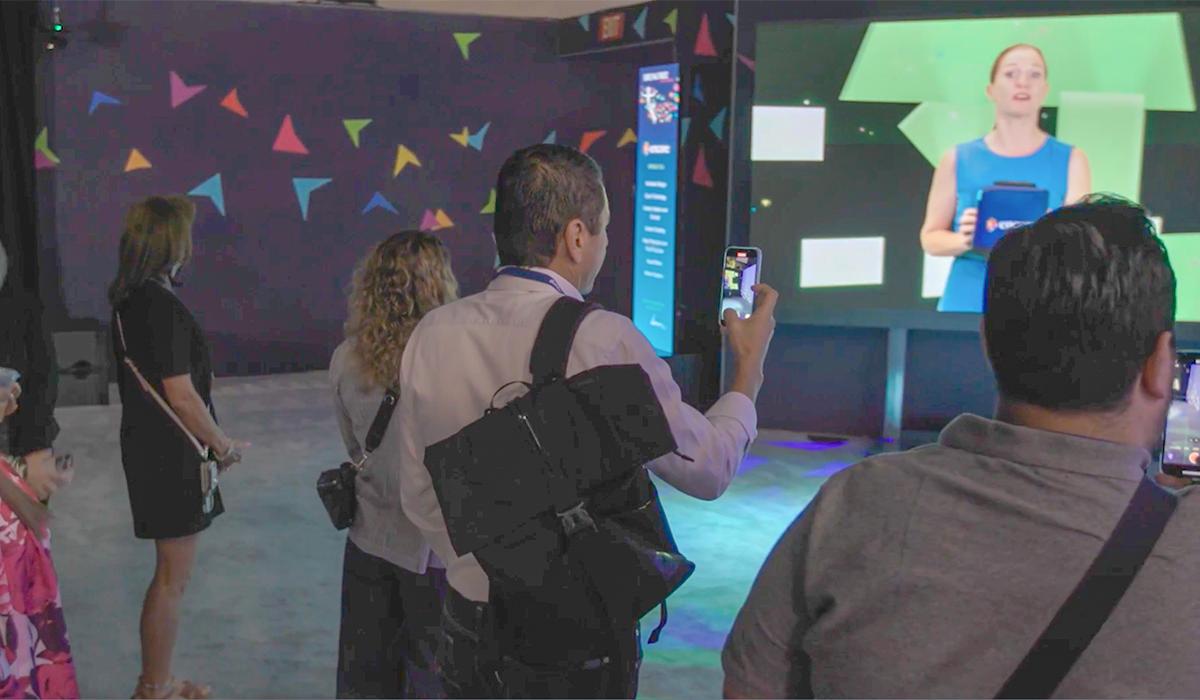 A group of people stands indoors, some filming with their phones, as they watch a large video screen showing a woman speaking in front of a colorful, geometric-patterned background.