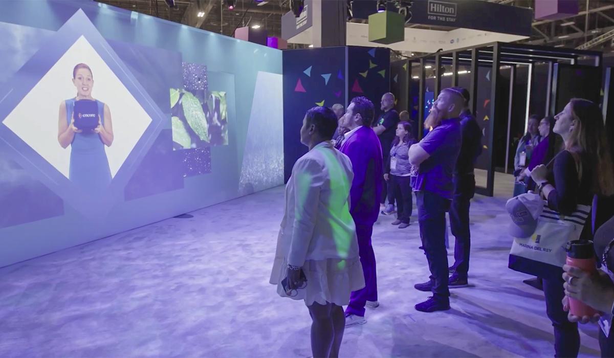 A group of people stands indoors watching a large digital screen displaying a woman speaking in a modern, tech-themed exhibit or event space.