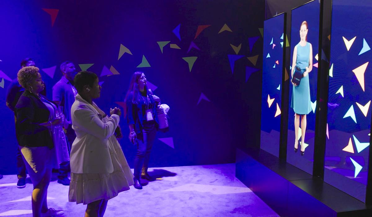 Four people stand in front of two large interactive screens displaying a woman in a blue dress, with colorful geometric shapes on a dark blue background, in an exhibit or event space with purple lighting.