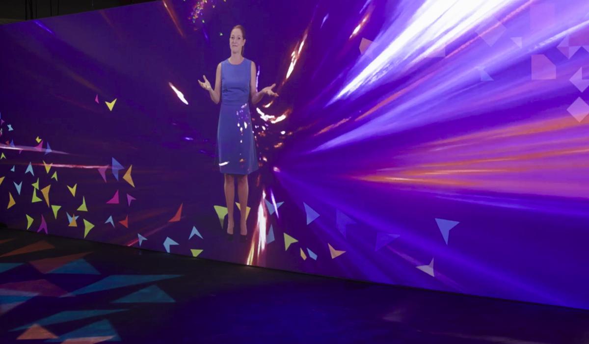 A woman in a blue dress gestures with her hands while standing in front of a vivid, abstract purple background with bright streaks and colorful triangular shapes.