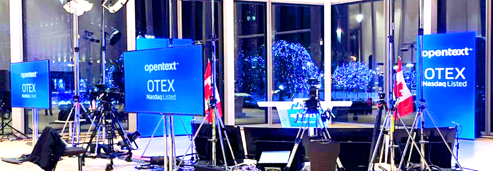 A brightly lit indoor event setup displays opentext OTEX Nasdaq Listed on screens, with cameras, lights, Canadian flags, and large windows showing blue-lit trees outside at night.