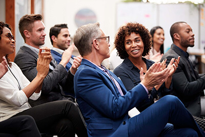 A diverse group of people sits in rows, smiling and applauding at an indoor event, suggesting they are attending a business presentation or conference.