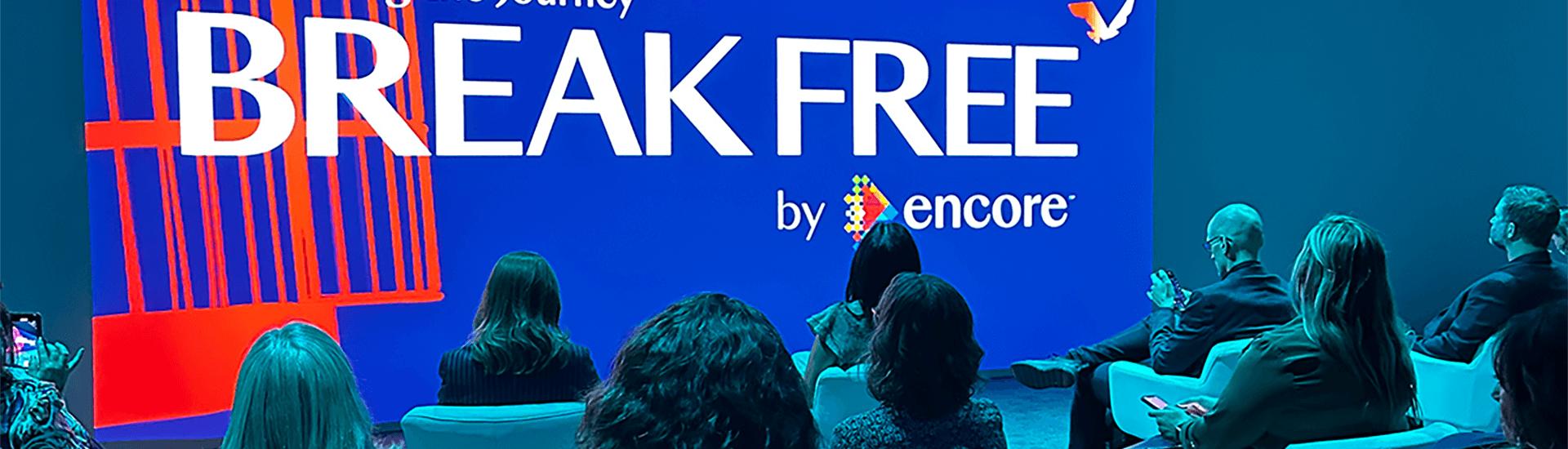 A group of people sit facing a large blue screen displaying BREAK FREE: IMEX America 2023 by Encore® with an orange illustration of a broken jail cell.