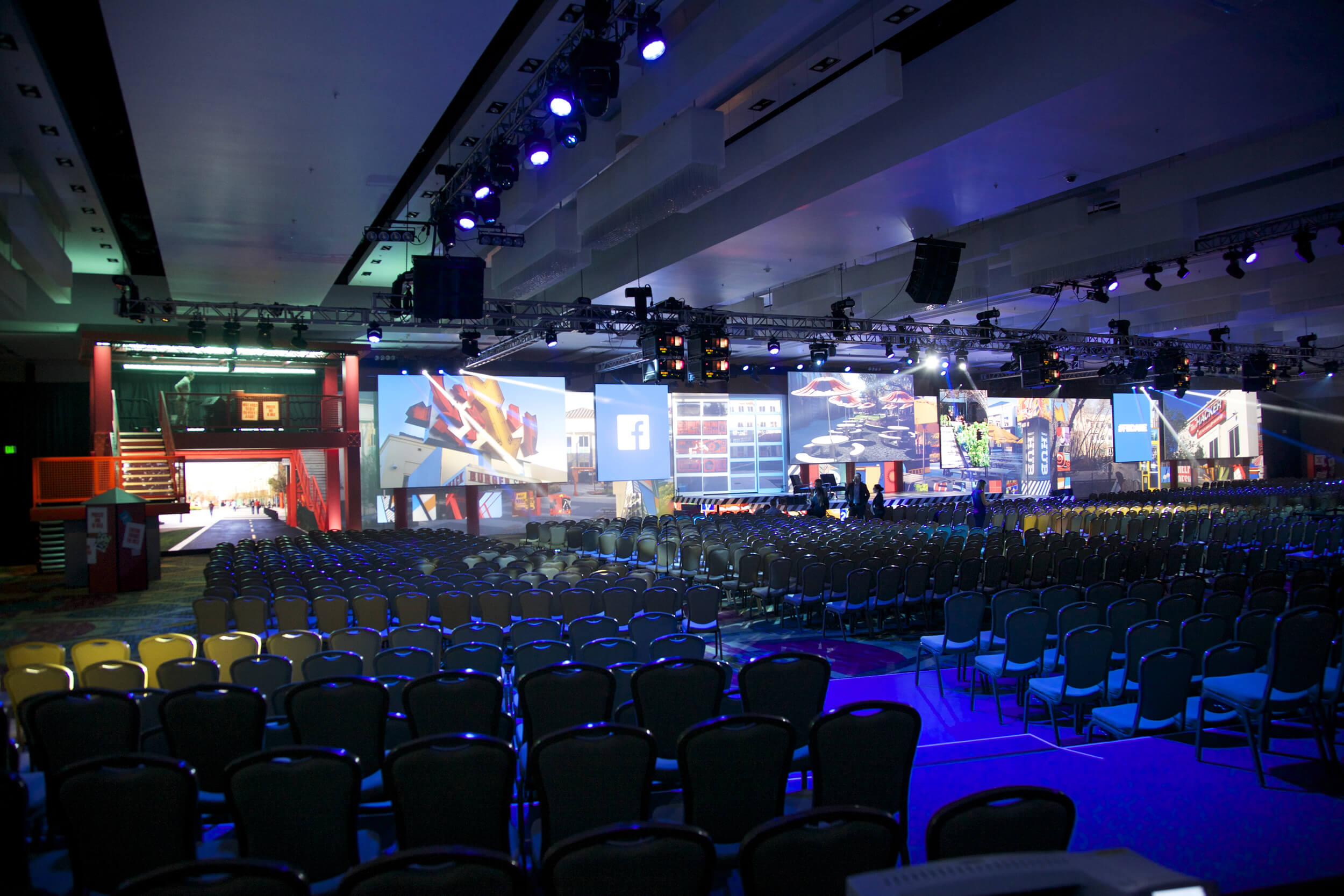 A large event hall filled with rows of empty chairs faces a stage with multiple projection screens showing vibrant images and social media logos, under blue and purple stage lighting.