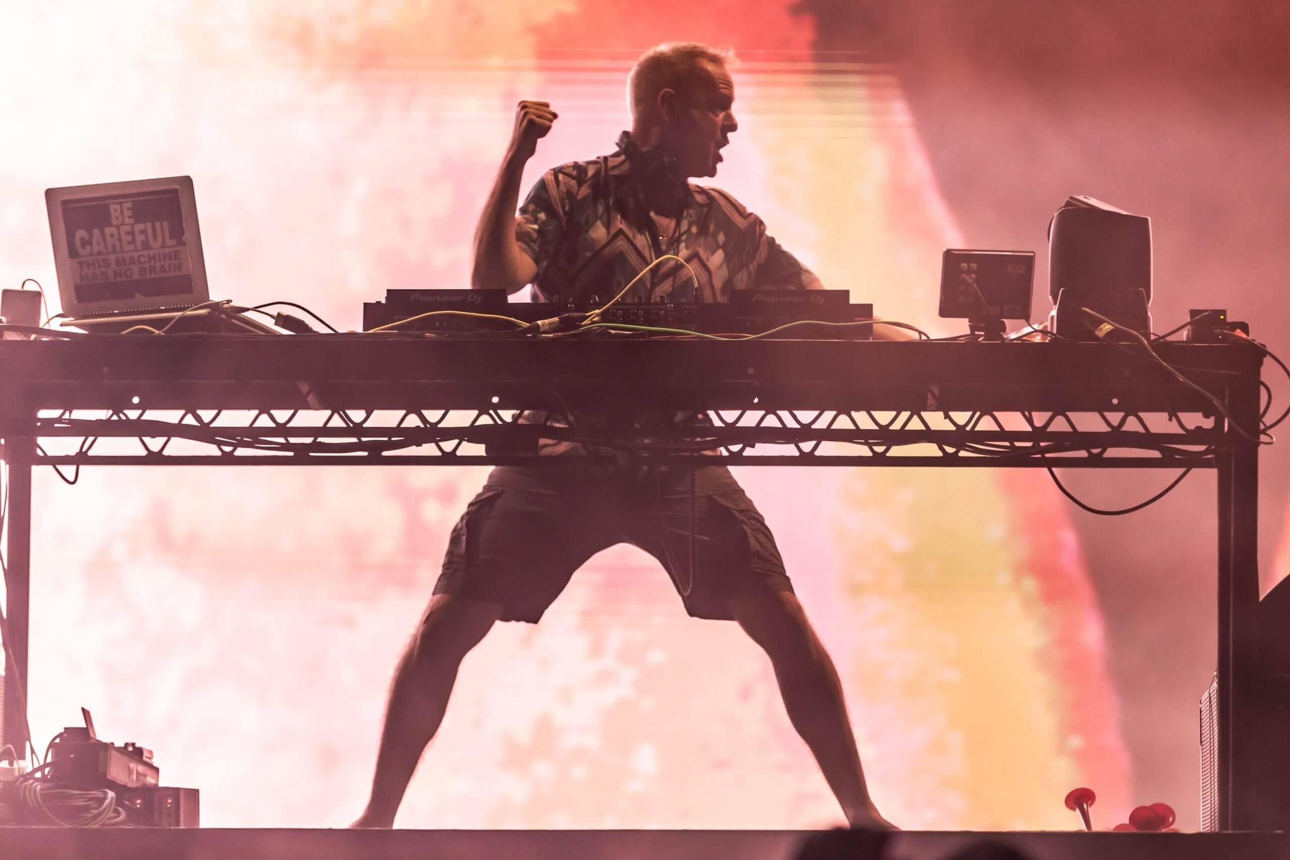 A DJ performs energetically on stage, standing behind a table with electronic equipment, silhouetted against a vibrant, colorful background.
