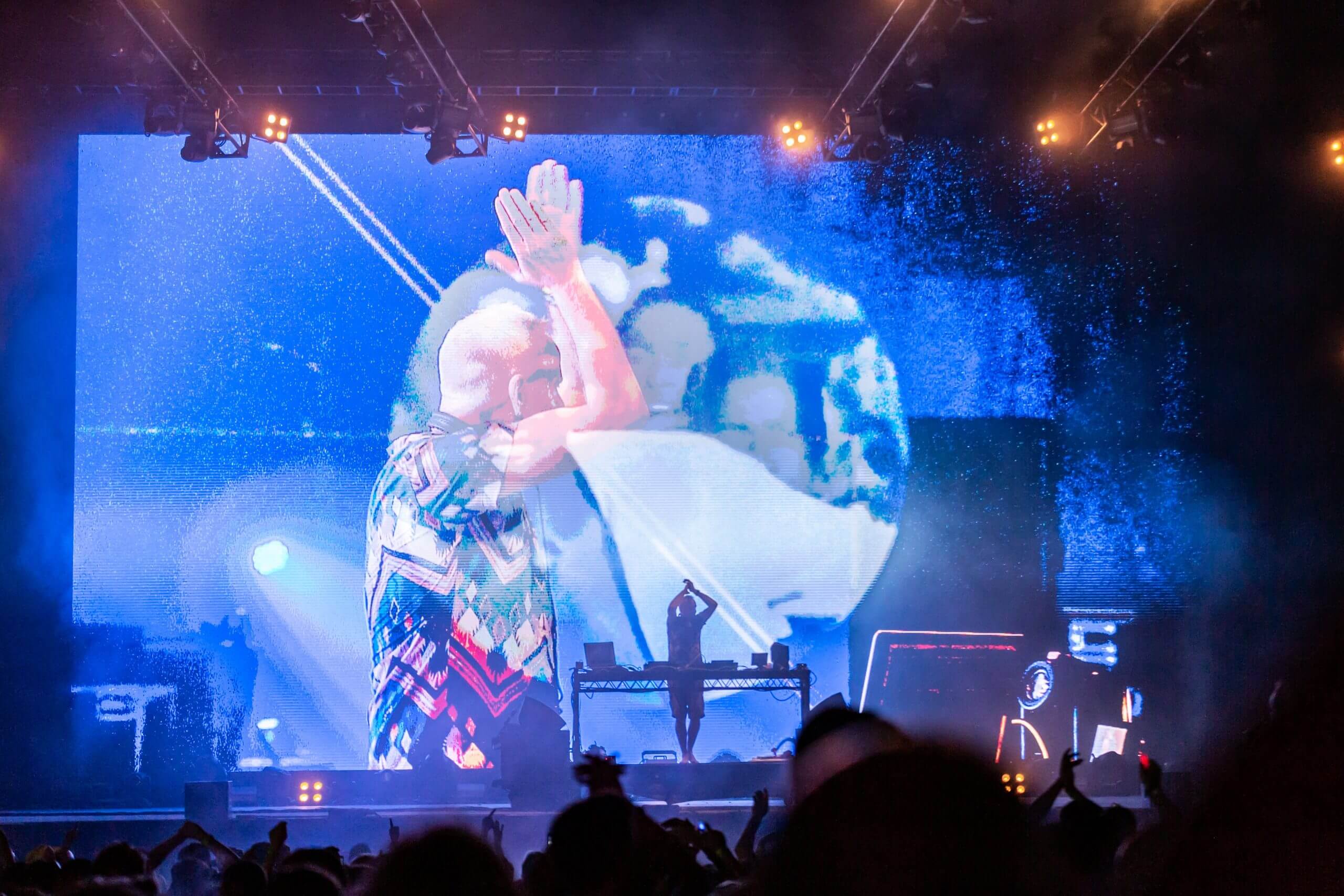 A DJ performs onstage in front of a large, vibrant screen displaying abstract graphics, with colorful lights and a cheering crowd in the foreground.