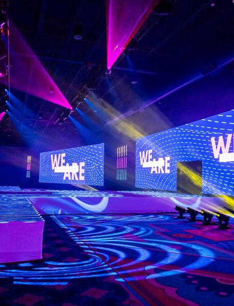 A vibrant event stage with large LED screens displaying WE ARE, colorful lighting, and patterned carpeted flooring in a spacious indoor venue.