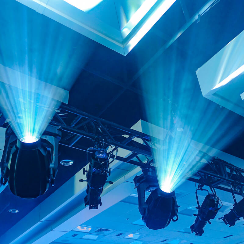 Stage lights mounted on a black truss project bright blue beams upward, illuminating the ceiling of an indoor venue with geometric ceiling panels.