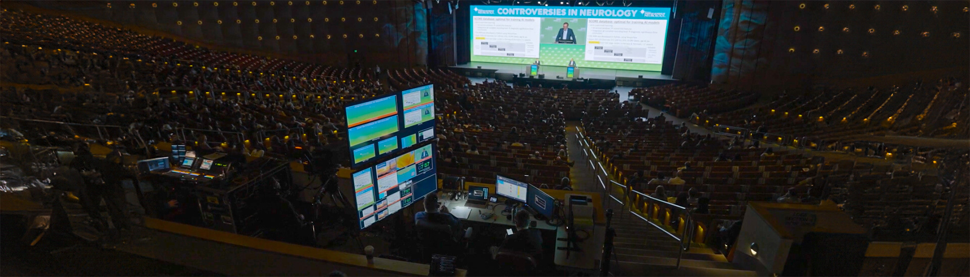 A large, dimly lit auditorium filled with people attending a conference. Multiple screens and control equipment are visible in the foreground, while two speakers stand on a stage beneath a brightly lit presentation screen.