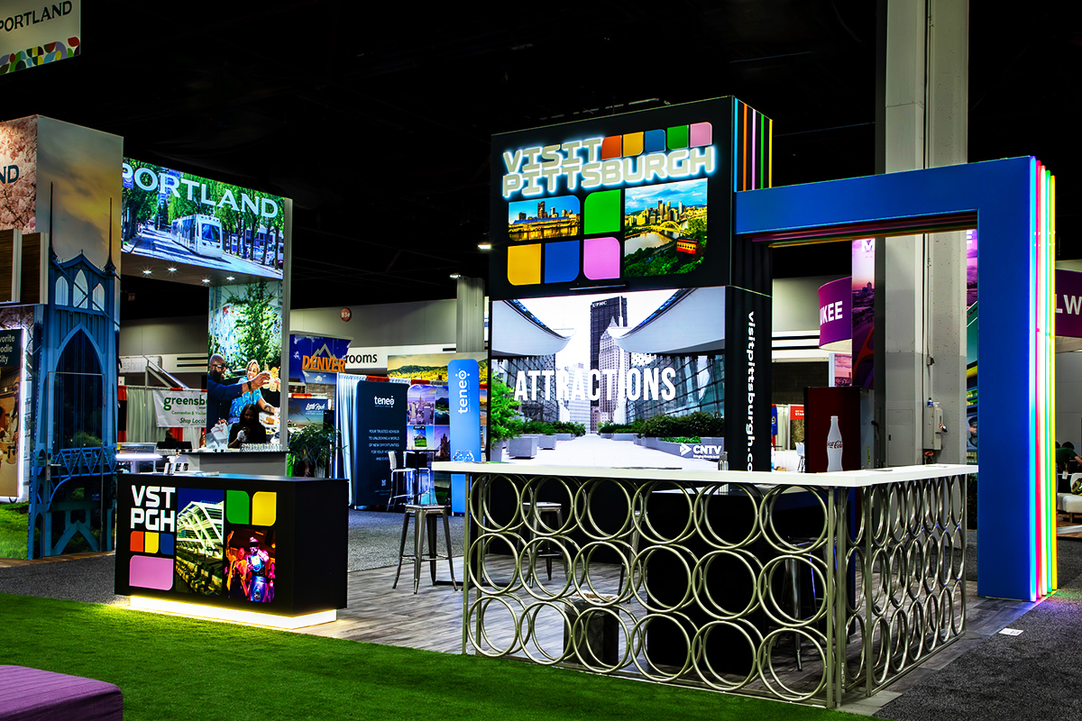 A colorful convention booth displays signs reading Visit Pittsburgh with vibrant city images, circular metal table, and digital screens showcasing local attractions, all set in a large exhibition hall.