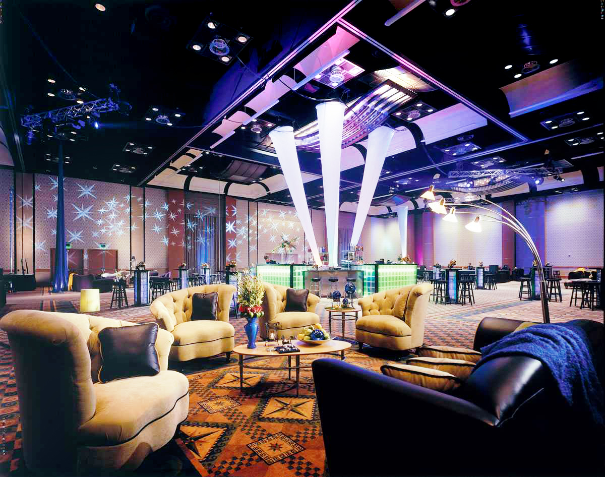 A stylish lounge with plush seating, modern decor, and a central illuminated bar under unique white light columns. Star-shaped lights are projected on the walls, creating a vibrant, upscale atmosphere.