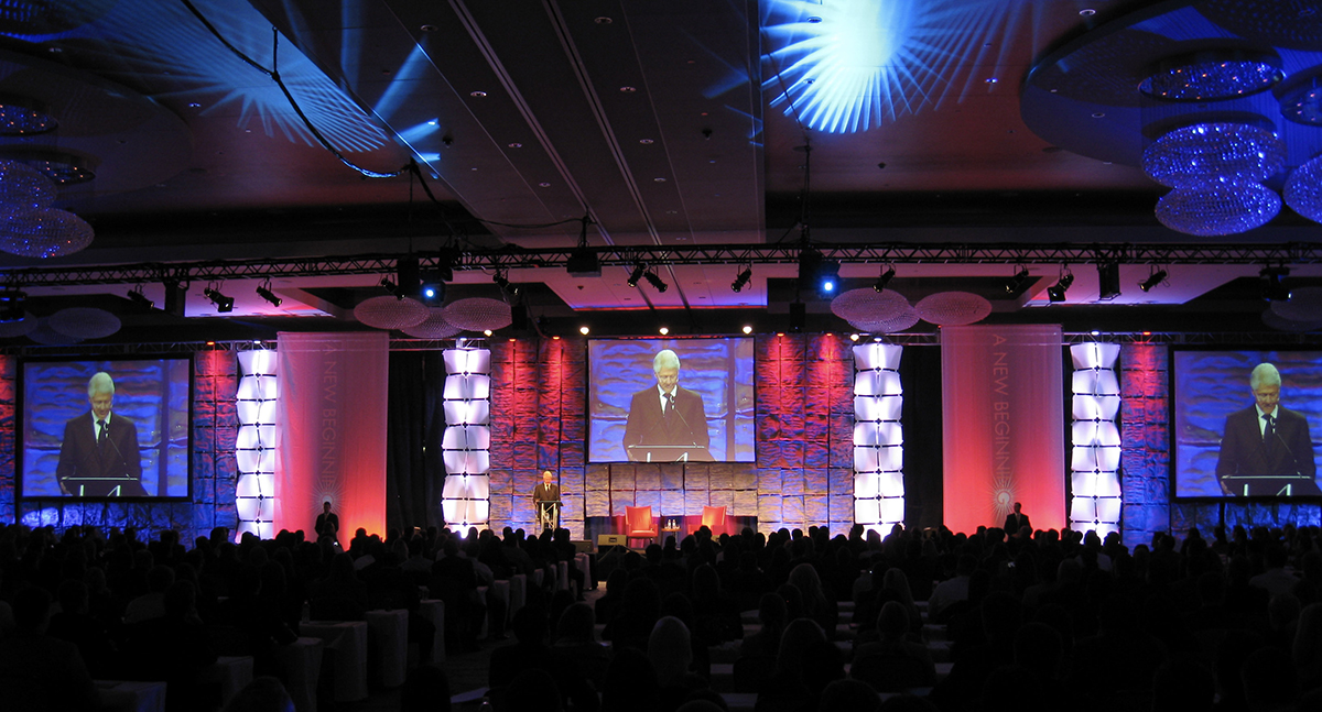 A large conference room with an audience facing a stage, where a person speaks at a podium. Two large screens display the speaker, and colorful lights illuminate the stage with blue and purple tones.