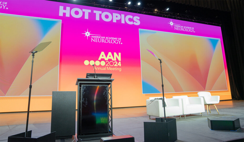 A brightly lit stage set for the AAN 2024 Annual Meeting, featuring a podium, teleprompters, white chairs, and large screens displaying HOT TOPICS and the American Academy of Neurology logo.