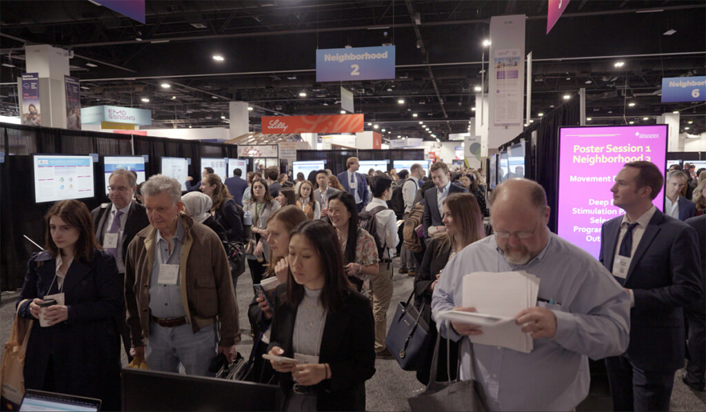 A large group of people attends a busy conference or convention, standing and walking between display screens and informational booths under signs and banners in a well-lit indoor venue.