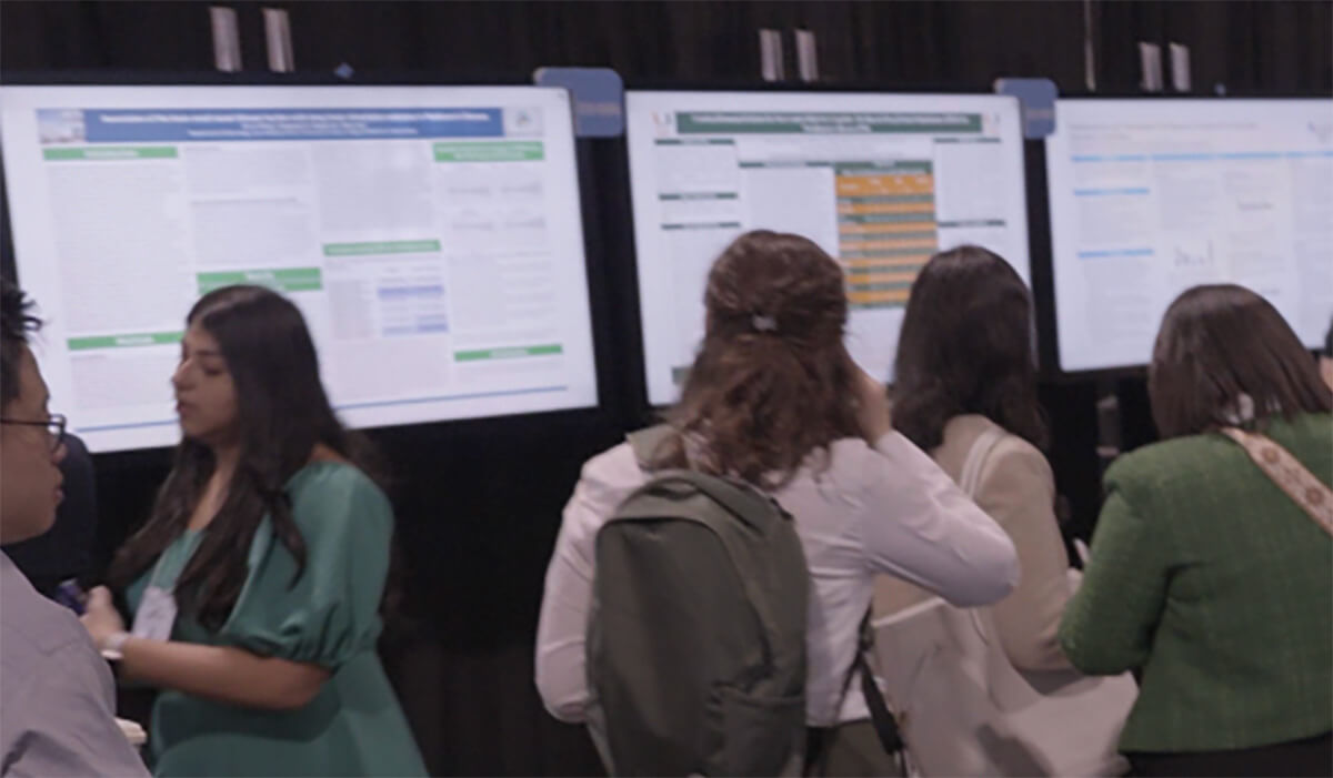 Several people stand in front of large, brightly lit presentation boards at an event, reading and discussing the displayed information. The scene appears busy and academic.