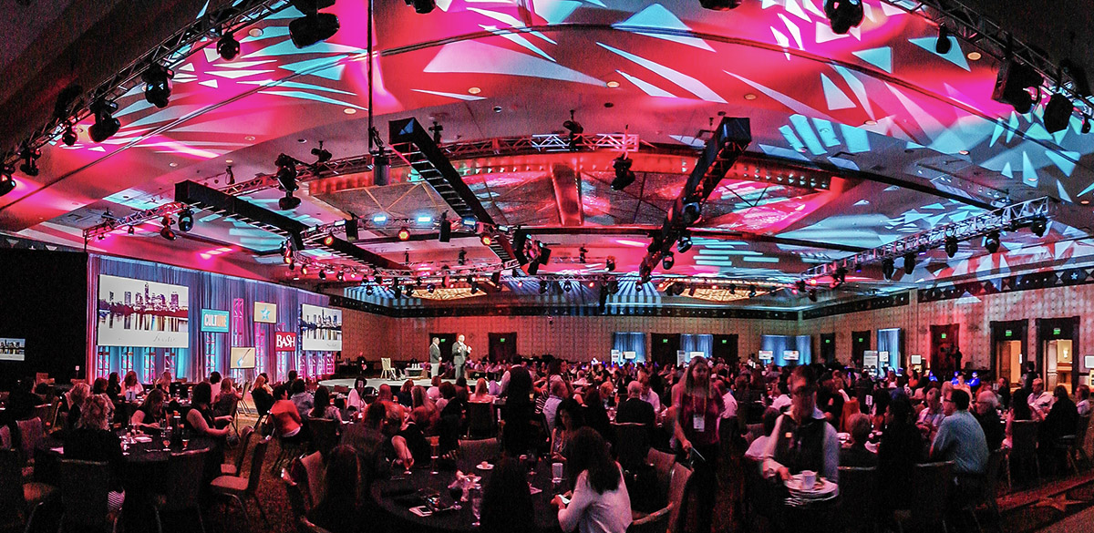 A large ballroom filled with seated people watching a stage presentation, with colorful pink and blue lighting patterns projected on the ceiling and walls, creating a vibrant atmosphere.
