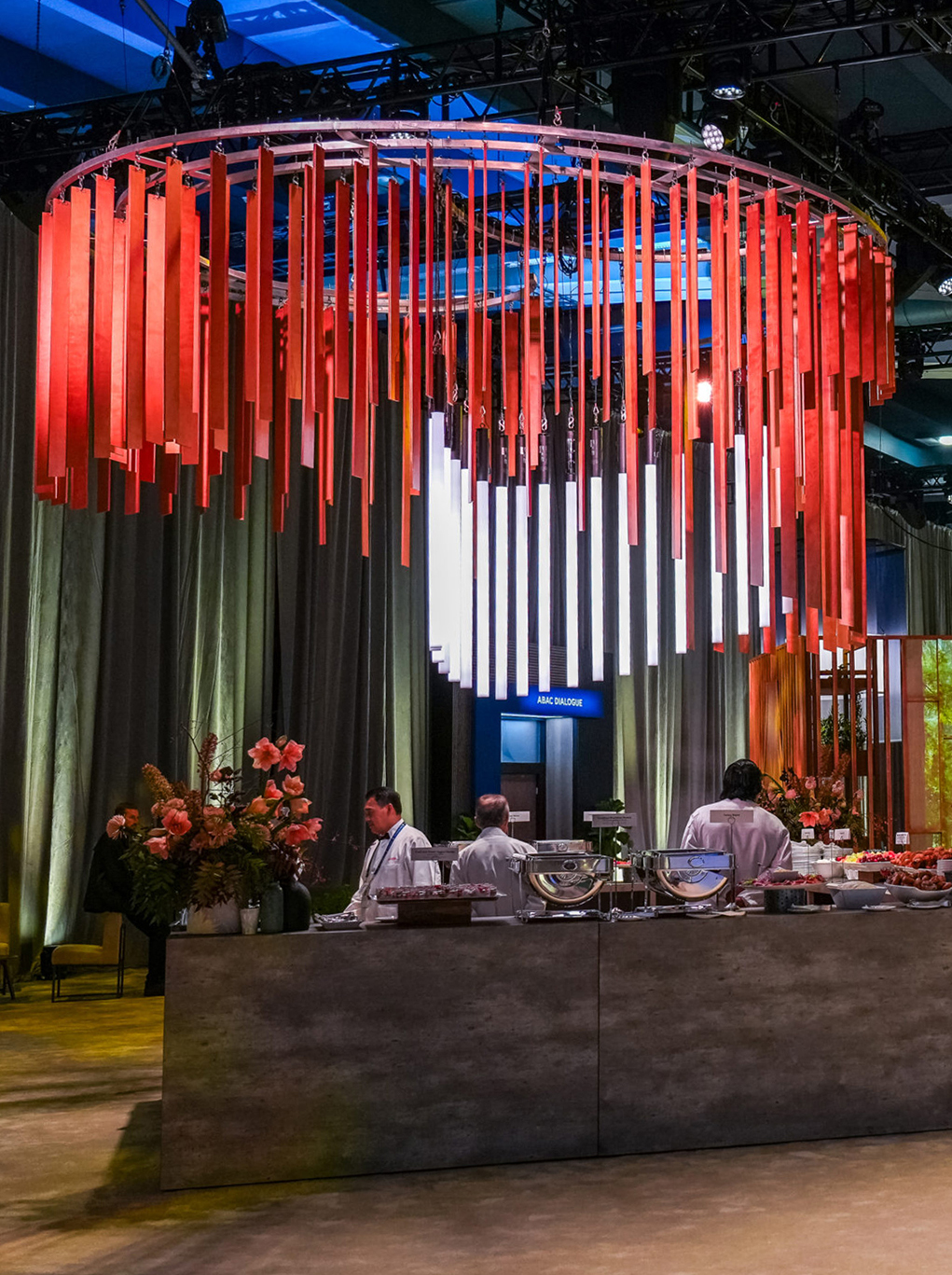 A modern buffet setup with chefs behind a stone counter, decorative flowers, and a large circular hanging light fixture with red and white vertical rods. The background features draped curtains and soft lighting.