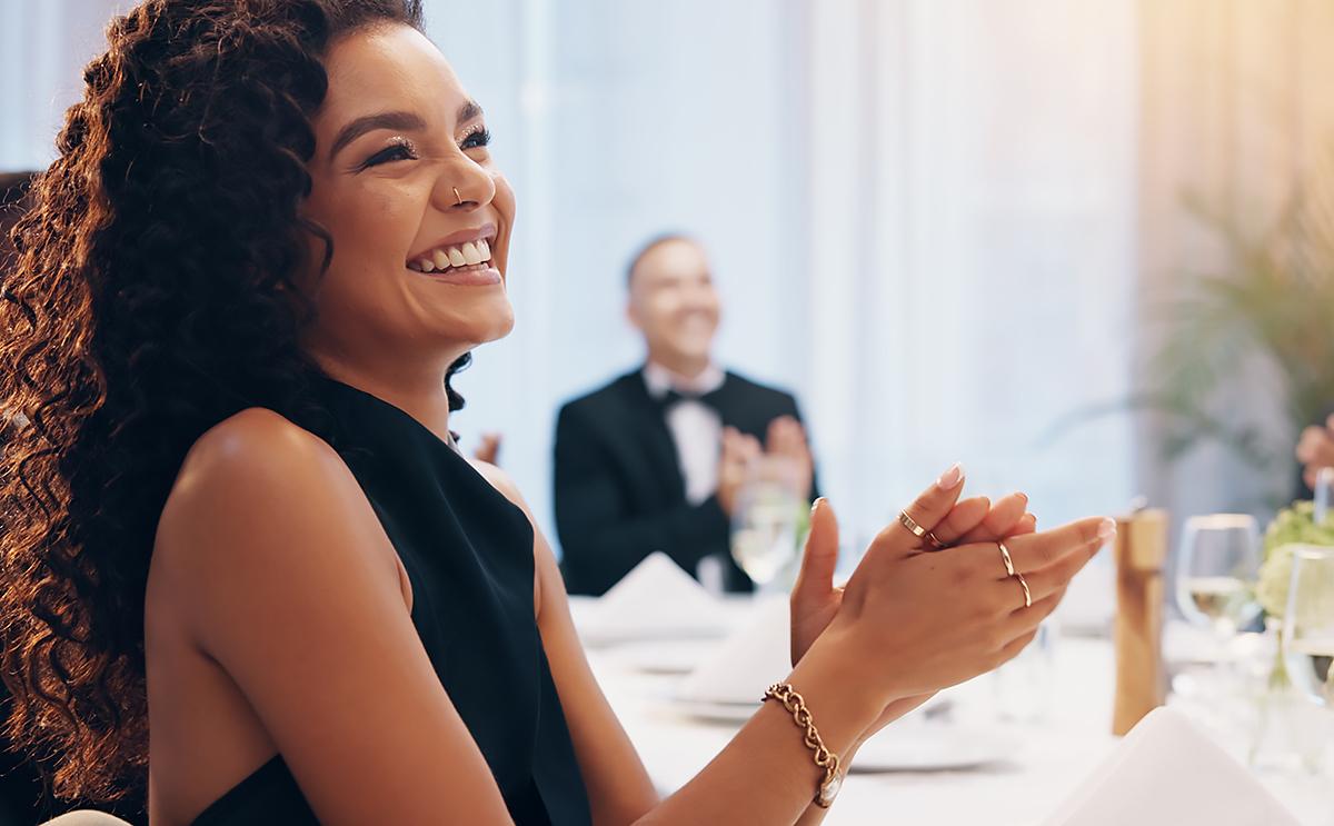 A woman with curly hair, wearing a sleeveless black dress and jewelry, smiles and claps at a formal event, with elegantly set tables and other guests in the background.