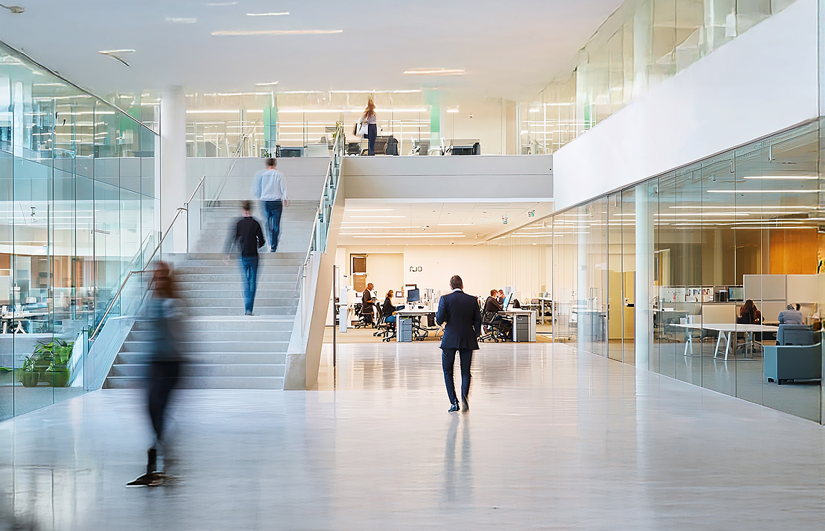 Modern office space with glass walls, open workstations, people walking and working, and a staircase leading to an upper level. The atmosphere is bright with natural and artificial lighting.