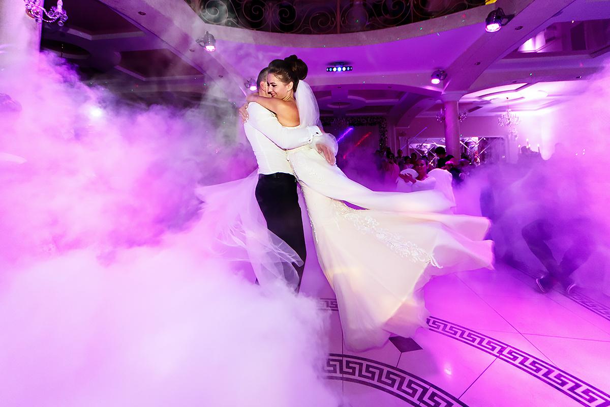 A bride and groom joyfully embrace and dance surrounded by colorful lights and dramatic fog on a decorated ballroom floor at their wedding reception.