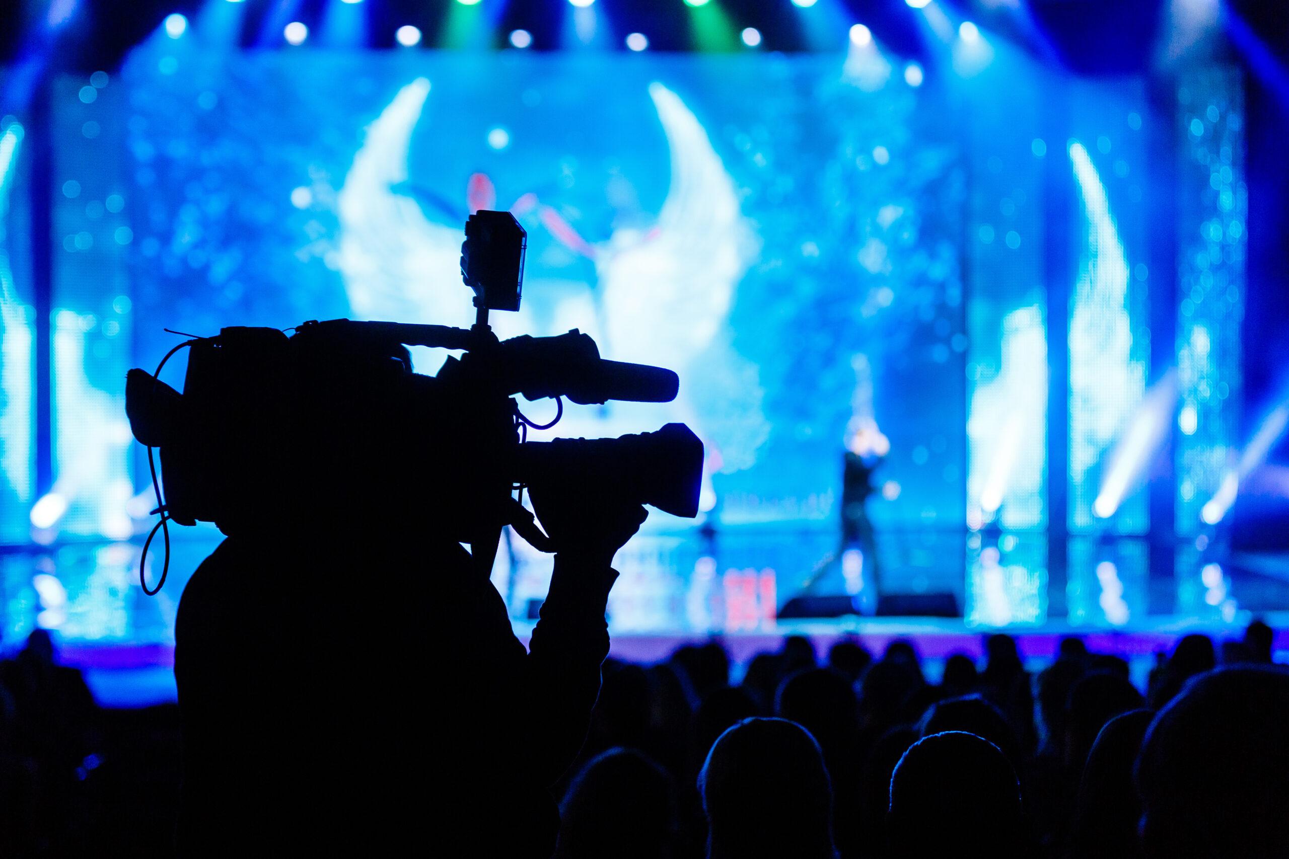 A silhouette of a camera operator filming a brightly lit stage with blue lighting and a blurred performer in the background, in front of an audience.