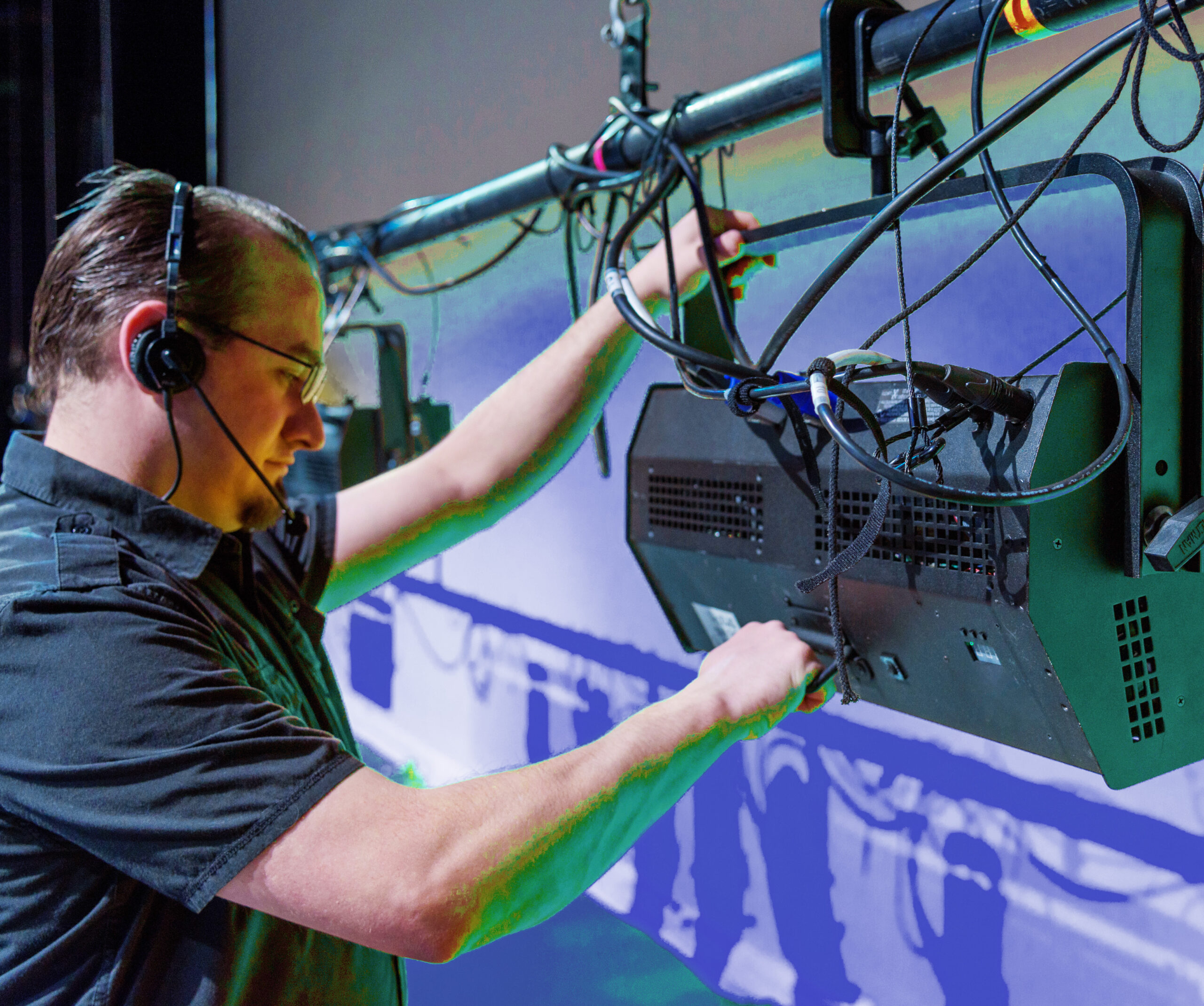 A technician wearing a headset adjusts a stage light fixture mounted on a rig, with cables visible around him and a blue-toned backdrop in the background.