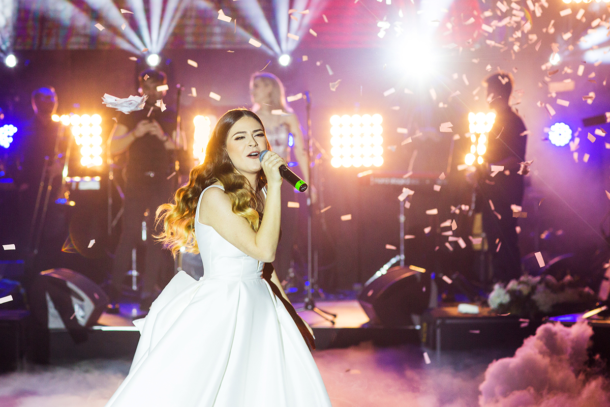 A woman in a white dress sings on stage under bright lights, surrounded by confetti, with a band playing in the background.