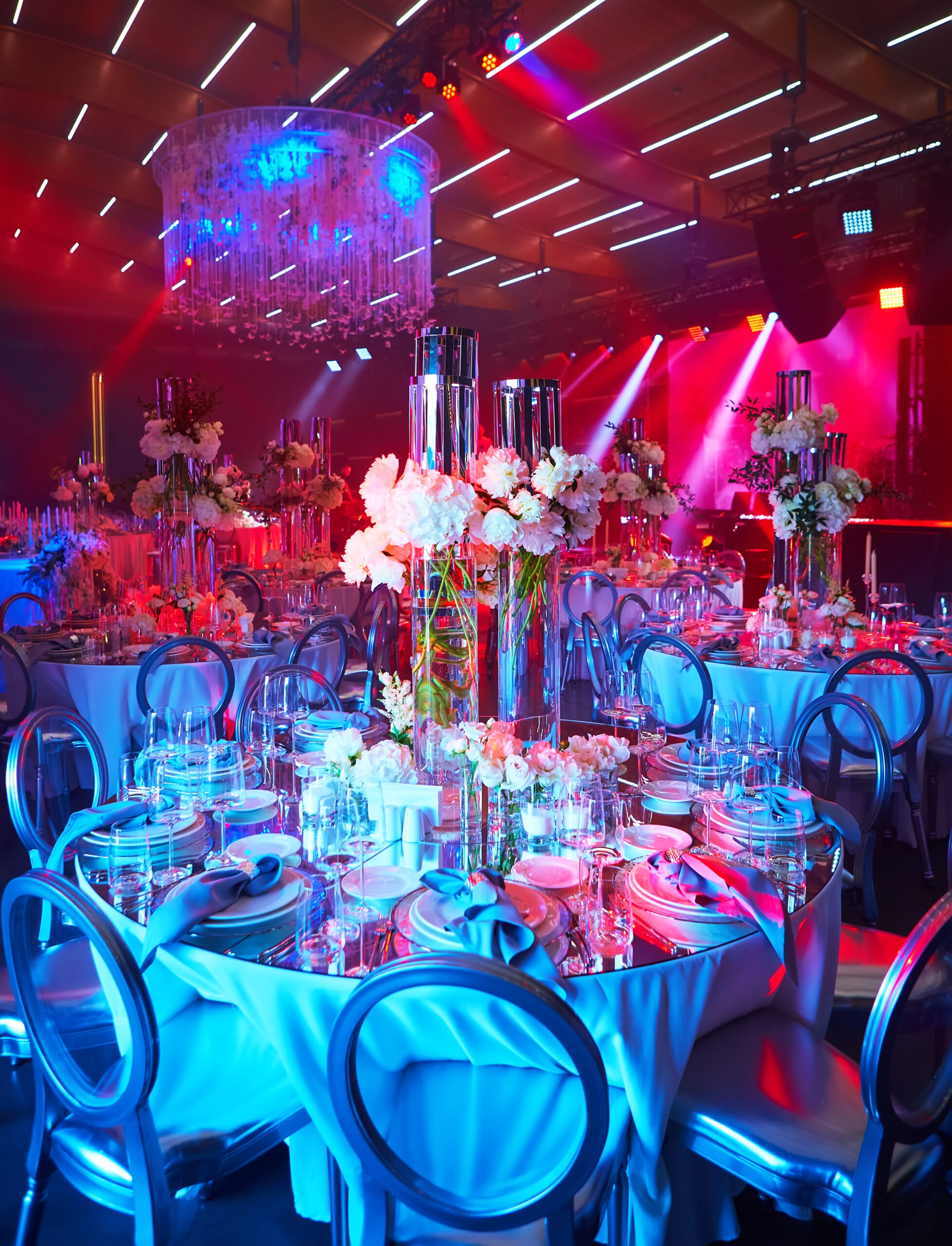 A lavish event hall with round tables set for dinner, featuring elegant glassware, plates, and tall floral centerpieces. Blue and red mood lighting and a large chandelier create a festive, upscale atmosphere.
