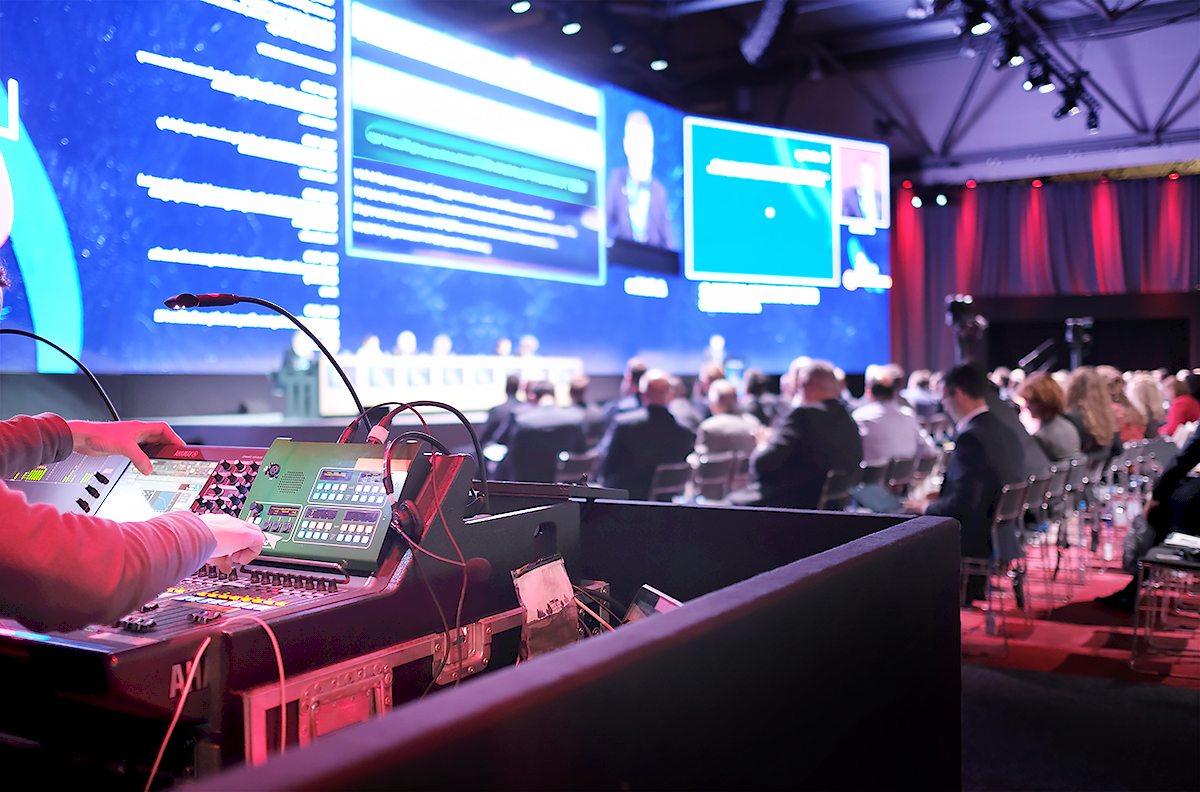 A person operates sound and lighting equipment at the back of a large conference hall, where attendees face a brightly lit stage with speakers and large presentation screens.