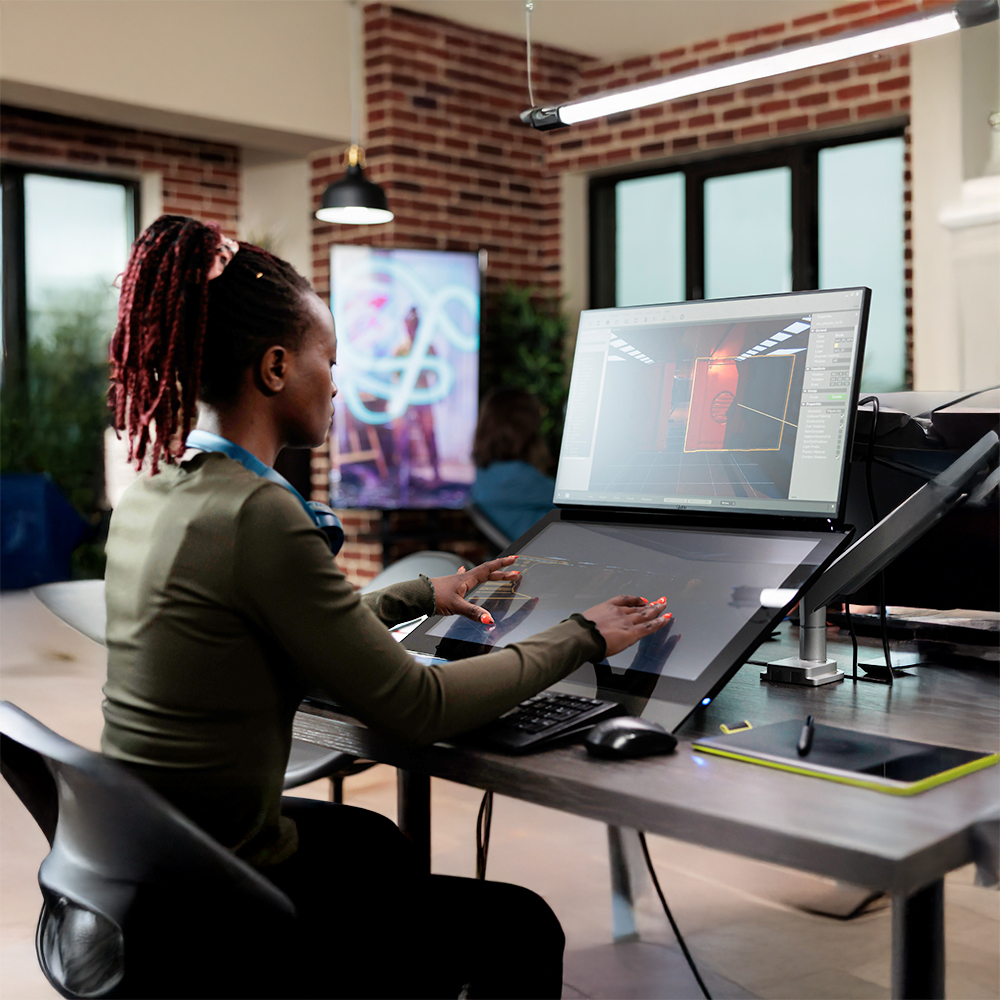 A person works at a large touchscreen computer, creating a 3D design of a red vending machine in a modern office with exposed brick walls and large windows.