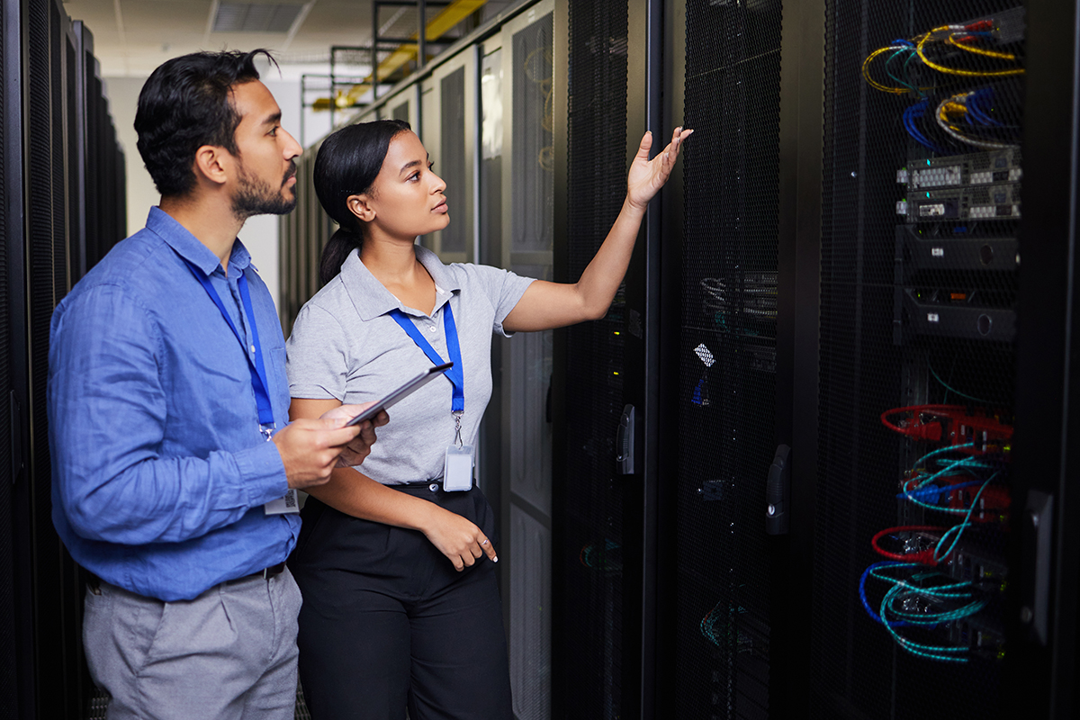 Two IT professionals, wearing ID badges, stand in a server room. One holds a tablet while the other gestures toward server racks filled with colorful cables and equipment.